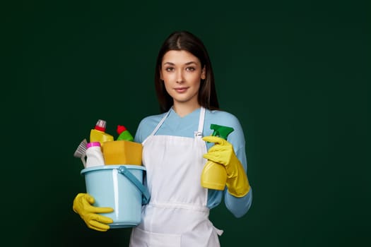 beautiful woman in yellow rubber gloves and cleaner apron holding bucket with cleaning supplies and detergent sprayer on dark green background.