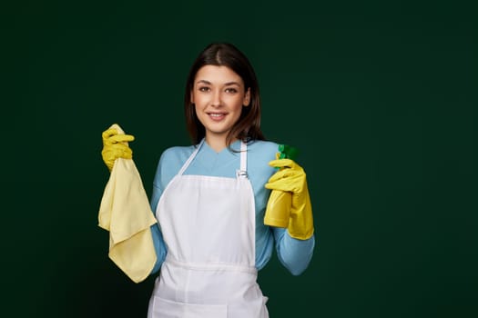 happy woman in yellow rubber gloves and cleaner apron with cleaning rag and detergent sprayer on dark green background.