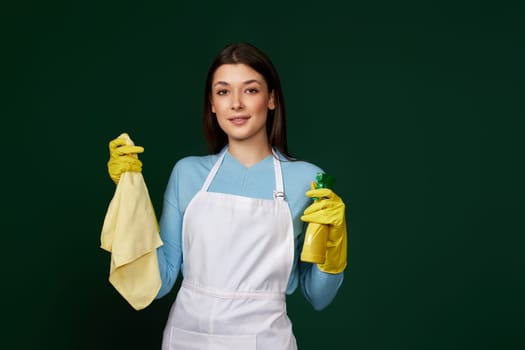 caucasian woman in yellow rubber gloves and cleaner apron with cleaning rag and detergent sprayer on dark green background.