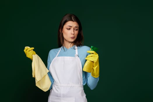 skeptic and nervous, frowning woman in gloves and cleaner apron with cleaning rag and detergent sprayer on green background.