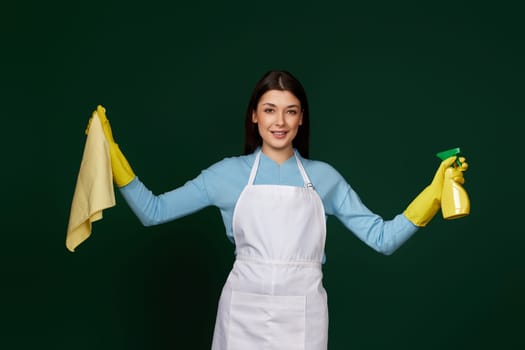 caucasian woman in yellow rubber gloves and cleaner apron with cleaning rag and detergent sprayer on dark green background.