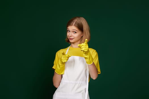 pretty blonde woman in yellow rubber gloves and cleaner apron pointing fingers to camera on green background.
