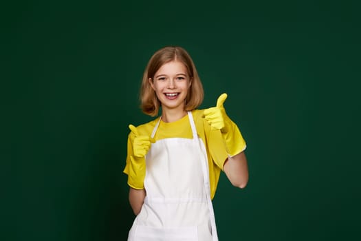 beautiful blonde woman in yellow rubber gloves and cleaner apron showing ok sign on green background.