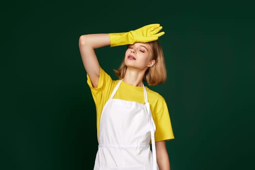 tired blonde woman in yellow gloves and cleaner apron on green background