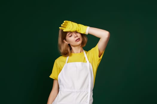 tired blonde woman in yellow gloves and cleaner apron on green background