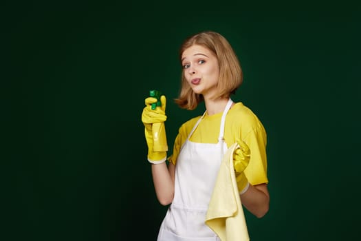beautiful caucasian blonde woman in yellow rubber gloves and cleaner apron with cleaning rag and detergent sprayer on dark green background.
