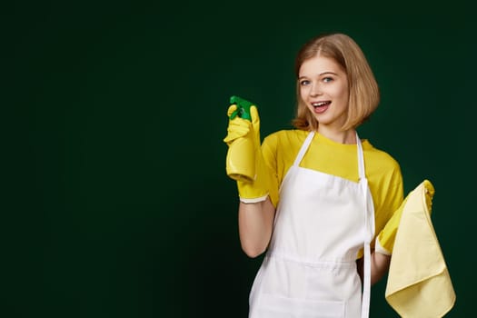 happy blonde woman in yellow rubber gloves and cleaner apron with cleaning rag and detergent sprayer on dark green background.