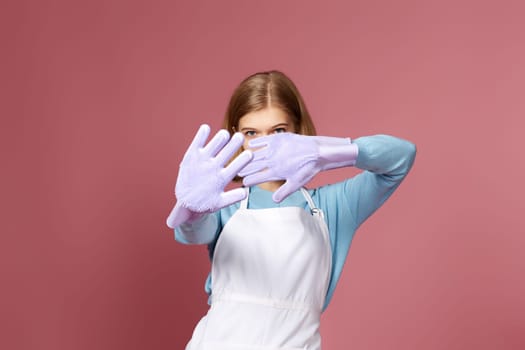 blonde girl in violet gloves and cleaner apron showing stop gesture on pink background. focus on hand