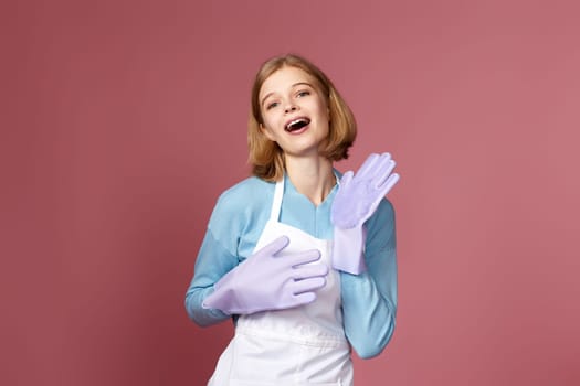 beautiful funny blonde woman in purple rubber gloves and cleaner apron dancing on pink background.