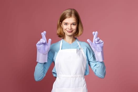 beautiful blonde caucasian girl in rubber gloves with finger crossed gesture on pink background.
