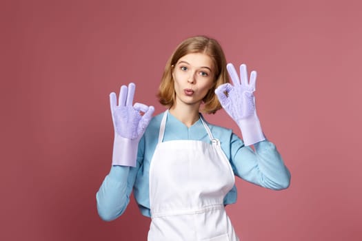 beautiful blonde woman in rubber gloves and cleaner apron showing ok sign on pink background.