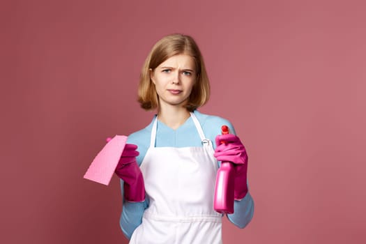 upset blonde woman in gloves and cleaner apron with cleaning rag and detergent sprayer on pink background.