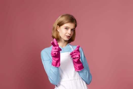 beautiful blonde woman in pink rubber gloves and cleaner apron pointing fingers to camera on studio background.