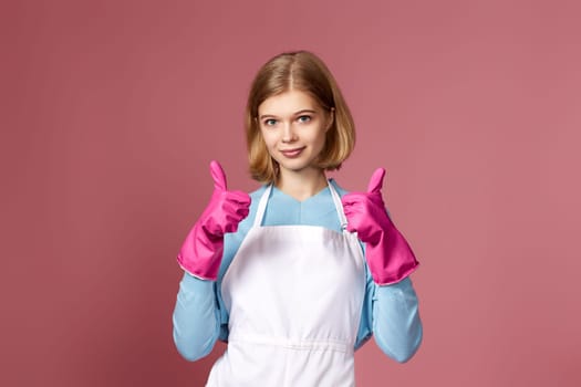 blonde woman in pink rubber gloves and cleaner apron showing thumbs up on pink background.