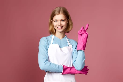 smiling blonde woman in rubber gloves and apron holding index finger up with new idea and looking at camera on pink background. cleaning