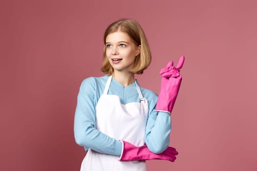 pretty smiling woman in rubber gloves and apron holding index finger up with new idea on pink background. cleaning