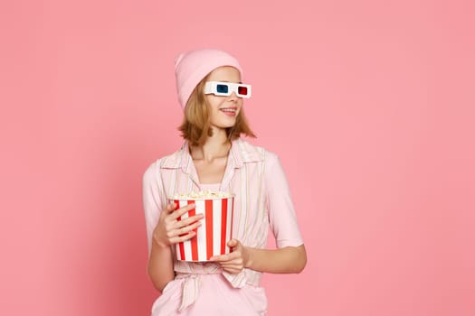 pretty blonde girl in pink t-shirt and hat holding bucket of popcorn and looking at side on pink background. copy space