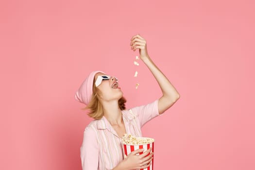pleased blonde woman in t-shirt and hat eating popcorn on pink background.