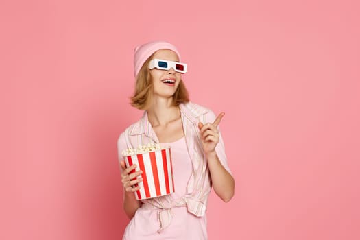 smiling woman in t-shirt and hat with popcorn pointing at the copy space on pink background.