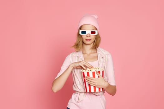 attractive blonde woman in 3d glasses holding bucket of popcorn isolated on pink background