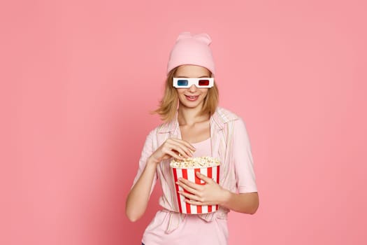 attractive blonde woman in 3d glasses holding bucket of popcorn isolated on pink background
