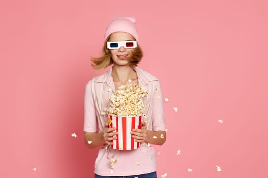 Cheerful blonde woman in 3d-glasses holding bucket of popcorn and jumping on pastel pink background.