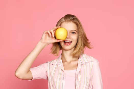 Portrait of smiling cheerful blonde woman covers one eye with fresh apple on pink background.