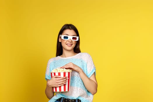 beautiful brunette girl in blue pullover watching movie film, holding bucket of popcorn isolated on yellow background