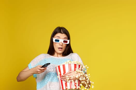 pretty woman with remote control holding bucket of popcorn and jumping on yellow background.