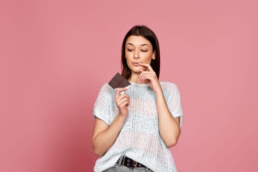 young woman in sweater holding delicious chocolate bar on pastel pink background