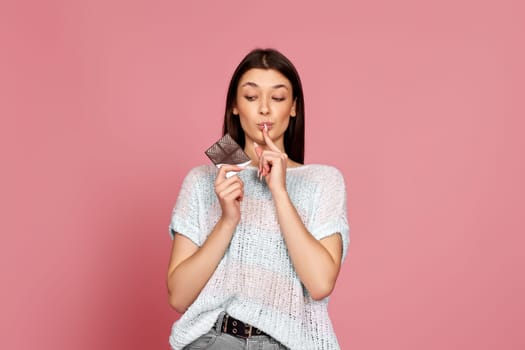 young woman in sweater holding delicious chocolate bar on pastel pink background