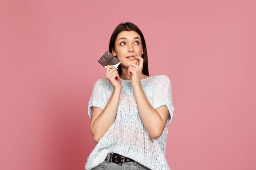 thoughtful brunette woman in sweater holding delicious chocolate bar on pastel pink background