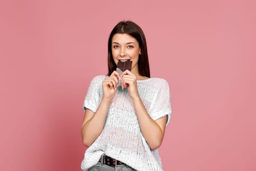 charming woman in sweater biting yummy sweet chocolate bar and looking at camera isolated on pastel pink background