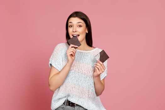 caucasian woman in sweater biting yummy chocolate bar and looking at camera on pastel pink background