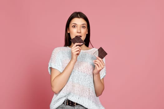 amazed woman in sweater biting sweet chocolate bar and looking at camera isolated on pastel pink background
