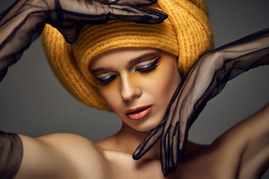 Beauty elegant blonde model girl with fashionable creative make-up, bright eye line wear yellow turban in black lace gloves on grey background.