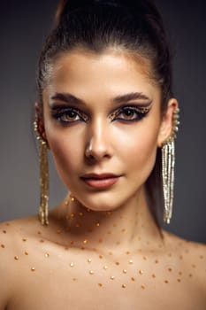 elegant fashionable model woman with creative eye makeup with arrows and rhinestones in long earring looking at the camera on gray background