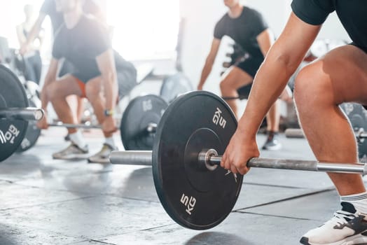 By using heavy weights. Group of young sportive people have crossfit day indoors in gym.