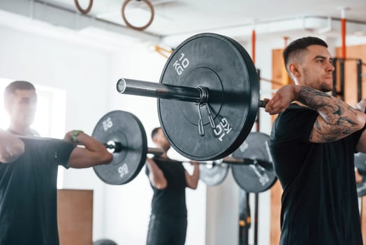By using heavy weights. Group of young sportive people have crossfit day indoors in gym.