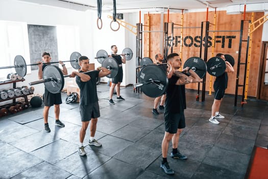 Together with barbells. Group of young sportive people have crossfit day indoors in gym.