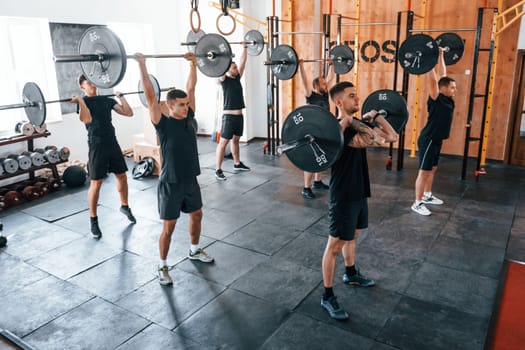 Together with barbells. Group of young sportive people have crossfit day indoors in gym.