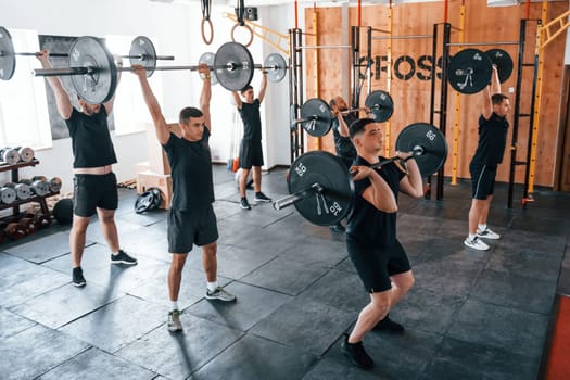 Together with barbells. Group of young sportive people have crossfit day indoors in gym.