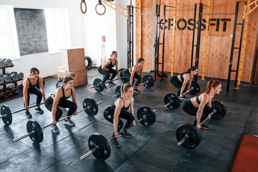 Together with barbells. Group of young sportive people have crossfit day indoors in gym.