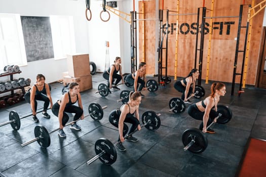 Together with barbells. Group of young sportive people have crossfit day indoors in gym.