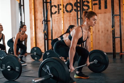 Together with barbells. Group of young sportive people have crossfit day indoors in gym.