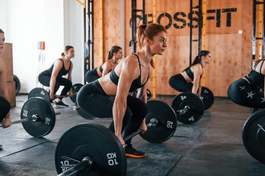 Together with barbells. Group of young sportive people have crossfit day indoors in gym.