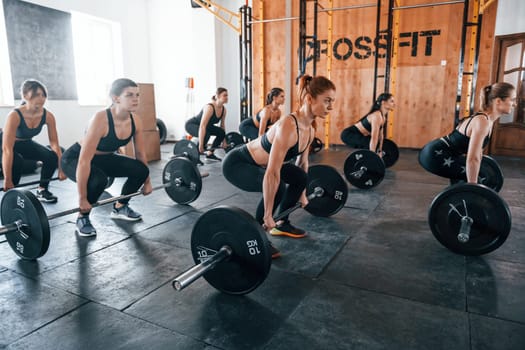 Together with barbells. Group of young sportive people have crossfit day indoors in gym.