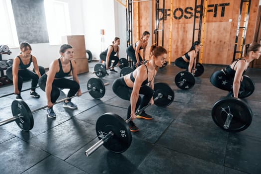 Together with barbells. Group of young sportive people have crossfit day indoors in gym.