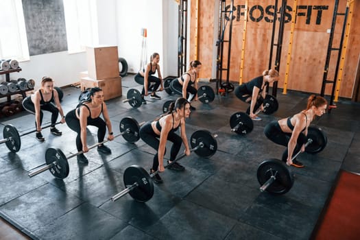 Together with barbells. Group of young sportive people have crossfit day indoors in gym.
