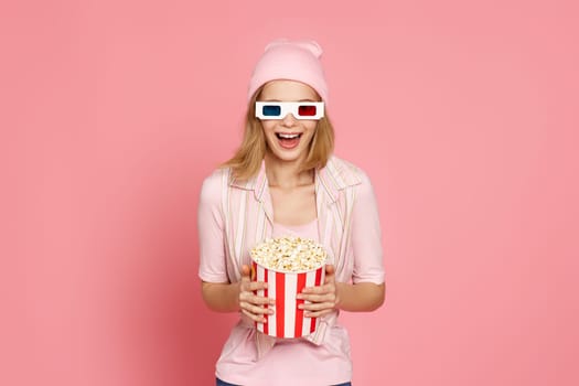 very excited young blonde woman holding bucket of popcorn on pastel pink background